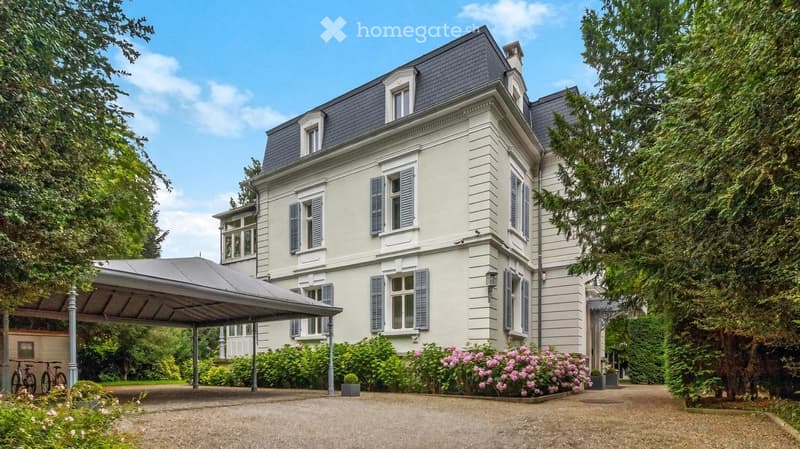A large, elegant two-story house with light beige walls, dark grey roof, multiple windows with shutters, and a covered driveway. The house is surrounded by lush greenery and flowering bushes.