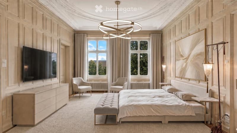 Elegant bedroom with a large bed, bench at the foot, two armchairs, dresser, television, and artwork on the wall. The room features large windows with curtains and a modern chandelier.