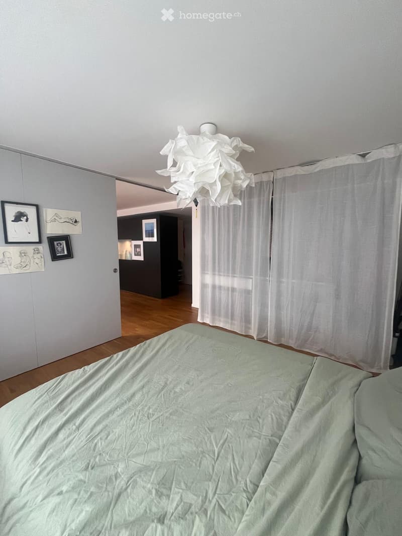 A modern bedroom with a neatly made bed, white curtains, and several picture frames on the wall. A hallway leads to another room with dark walls and additional artwork.