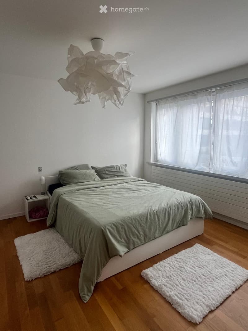 Modern bedroom with a large bed, nightstand, and two white rugs on a wooden floor. The room features a large window with white curtains and a decorative hanging light fixture.