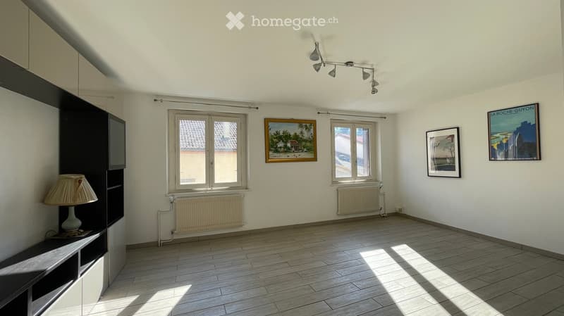 Empty room with light-colored walls, wooden flooring, and large windows. Features include built-in shelving, a lamp, and several framed pictures on the walls.