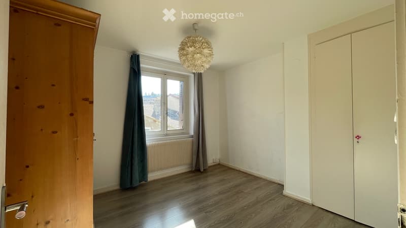 Empty room with wooden flooring, white walls, a window with green curtains, a radiator, and a hanging light fixture.