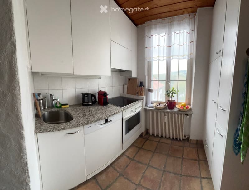 A compact kitchen with white cabinets, granite countertops, and a tiled floor. Features include a sink, oven, dishwasher, coffee maker, and a small window with a view of greenery.