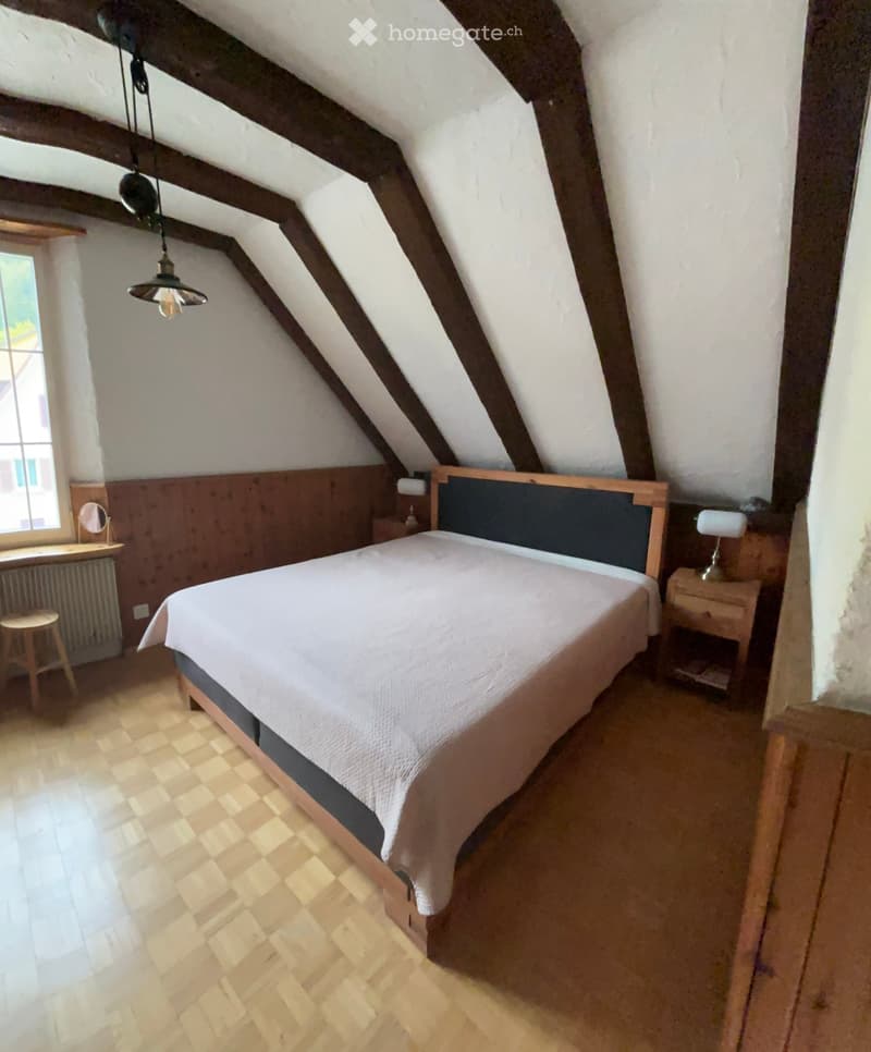Cozy attic bedroom with wooden slanted ceilings, a single bed with white bedding, a wooden nightstand with a lamp, a small wooden stool, and a mirror on the wall.