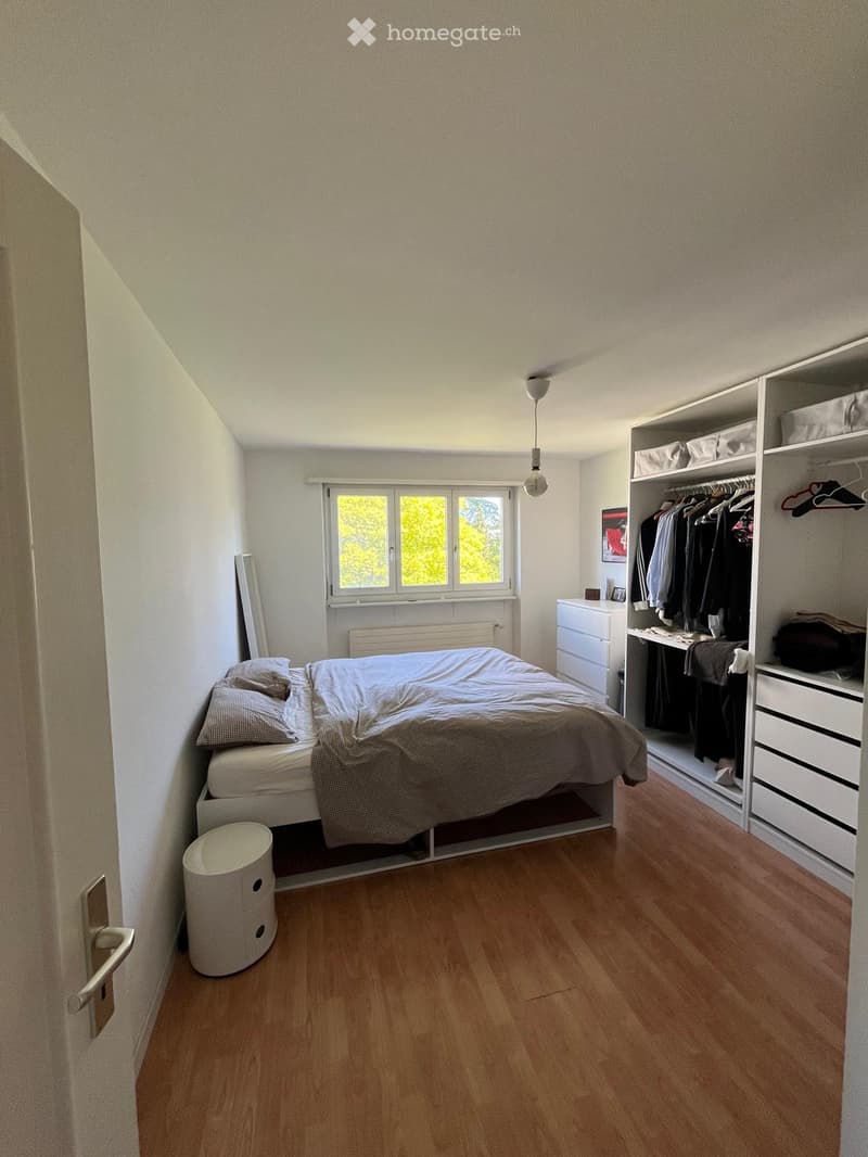 Cozy bedroom with wooden flooring, a bed with pillows and a blanket, a wardrobe with clothes, a small drawer, and a round stool near the door.