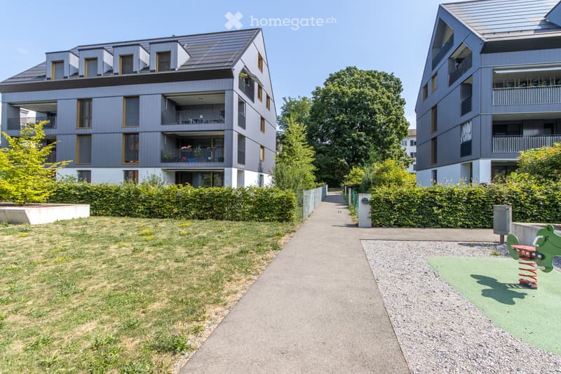Modern multi-story residential building with grey facade, balconies, and surrounding greenery. Pathway leads to a small playground area with a green spring toy.