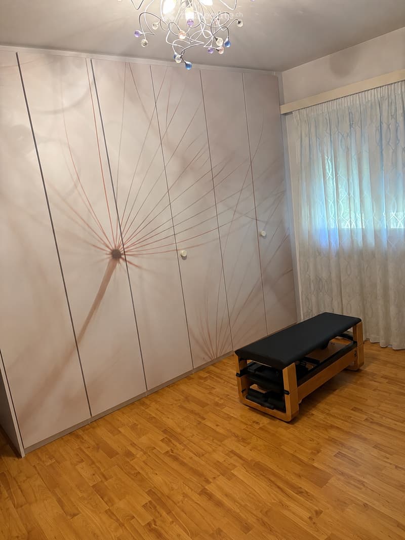 A clean, unfurnished room with wooden flooring, a massage table, and a large window with curtains.