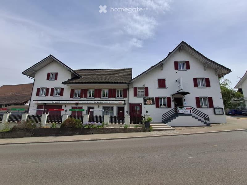 Two-story white building with red shutters and signage for "DA MARIO" and "KINDER PARADIES" visible. Features a staircase leading to an entrance, small garden area, and a parking area nearby.