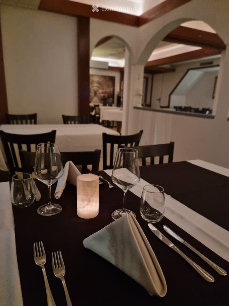 Elegantly set dining table with wine glasses, candle, napkins, and cutlery, surrounded by dark wooden chairs.