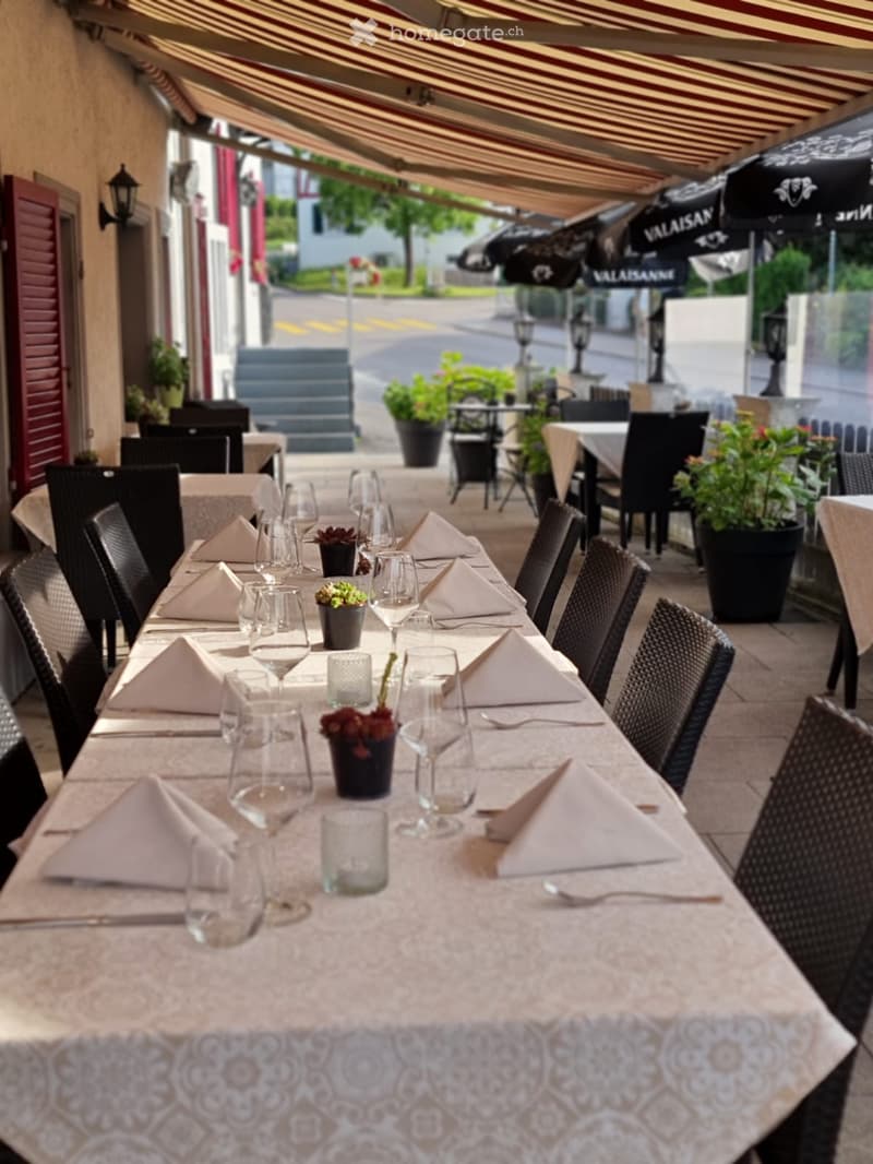 An outdoor dining area with a long table set for a meal, featuring wine glasses, napkins, and small flower pots as centerpieces. The area is covered by a striped awning and surrounded by additional tables and chairs.