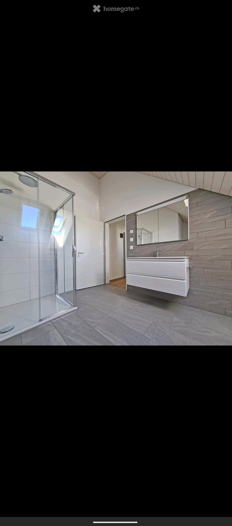Modern bathroom with a glass-enclosed shower, a white sink cabinet, and a large mirror above the sink. The walls are tiled, and the floor is covered with grey tiles.