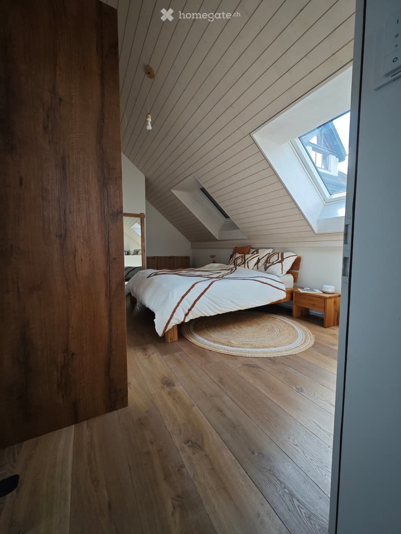 Cozy attic bedroom with wooden floors, slanted wooden ceiling, skylight windows, and a neatly made bed with pillows and a duvet.