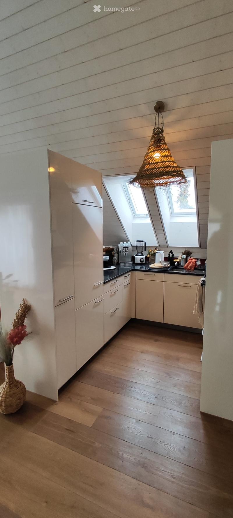 Modern kitchen with light-colored cabinets, wooden flooring, and a hanging pendant light. Appliances and a vase are visible on the counter.