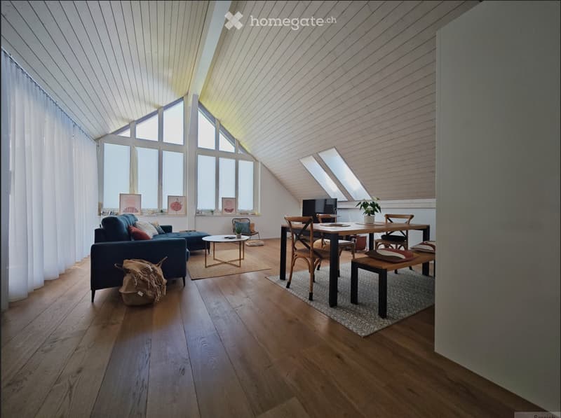 Bright, airy room with wooden floors, large windows, and skylights. Features a blue sofa, coffee table, dining table with chairs, and a small plant on a stool.