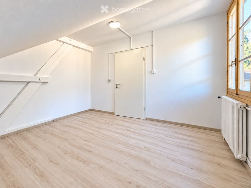 Empty attic room with wooden flooring, white walls, a single door, and a radiator near the window.