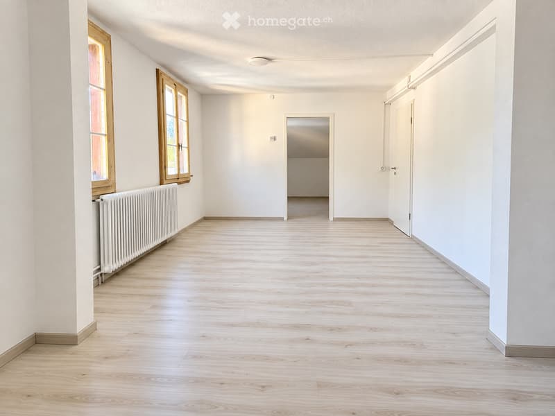 Empty hallway with light wooden flooring, white walls, and a radiator on the left side. There are two windows with wooden frames and a door at the end of the hallway.