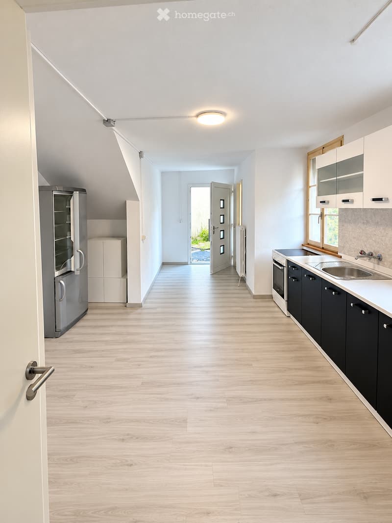 Bright, modern hallway with light wooden flooring, white walls, and recessed ceiling lighting. Features a built-in kitchen area with cabinets, sink, and appliances visible on the right side, and a refrigerator on the left. An open doorway leads to another