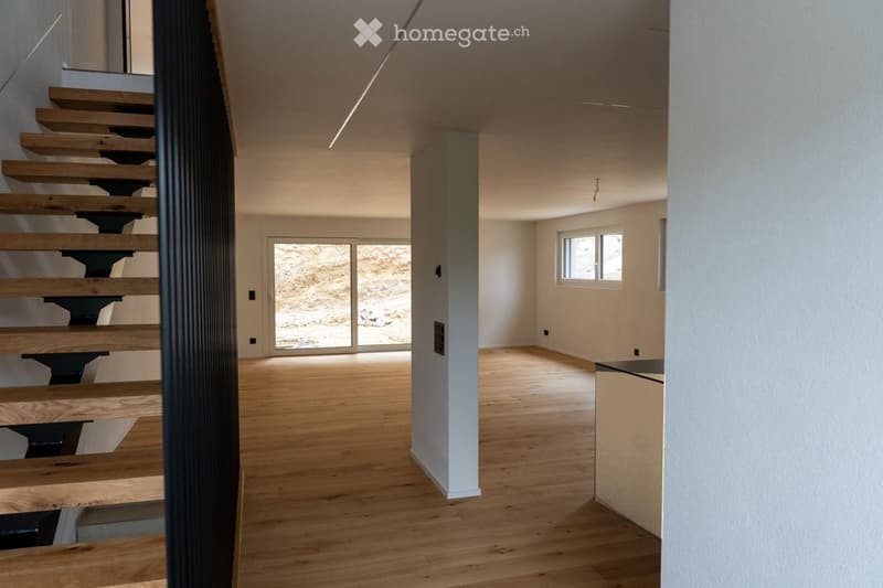 Empty room with wooden flooring, white walls, and a staircase leading upwards. The room has large windows and a door leading to an outdoor area.