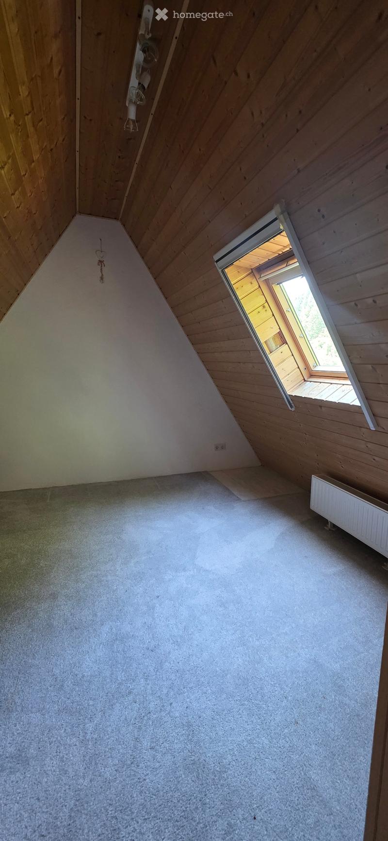 Empty attic room with wooden slanted ceilings, carpeted floor, and a small window opening to the outside.