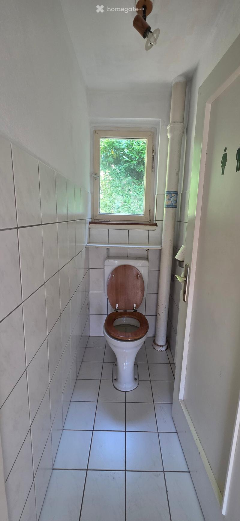 Small bathroom with white tiled walls and floor, a toilet with a wooden seat, a small window above the toilet, and a shelf above the window.