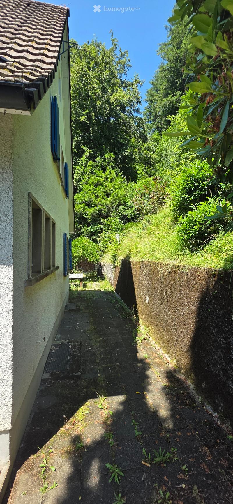 A narrow, paved pathway flanked by a stone wall and a building with a tiled roof, surrounded by greenery and trees.