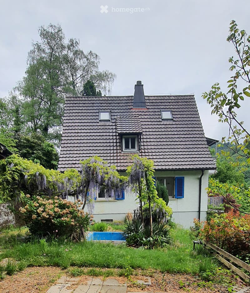 A two-story house with a gray tiled roof, white walls, and blue shutters. The house is surrounded by lush greenery, including trees, shrubs, and flowering plants. A small blue swimming pool is visible in the garden area.