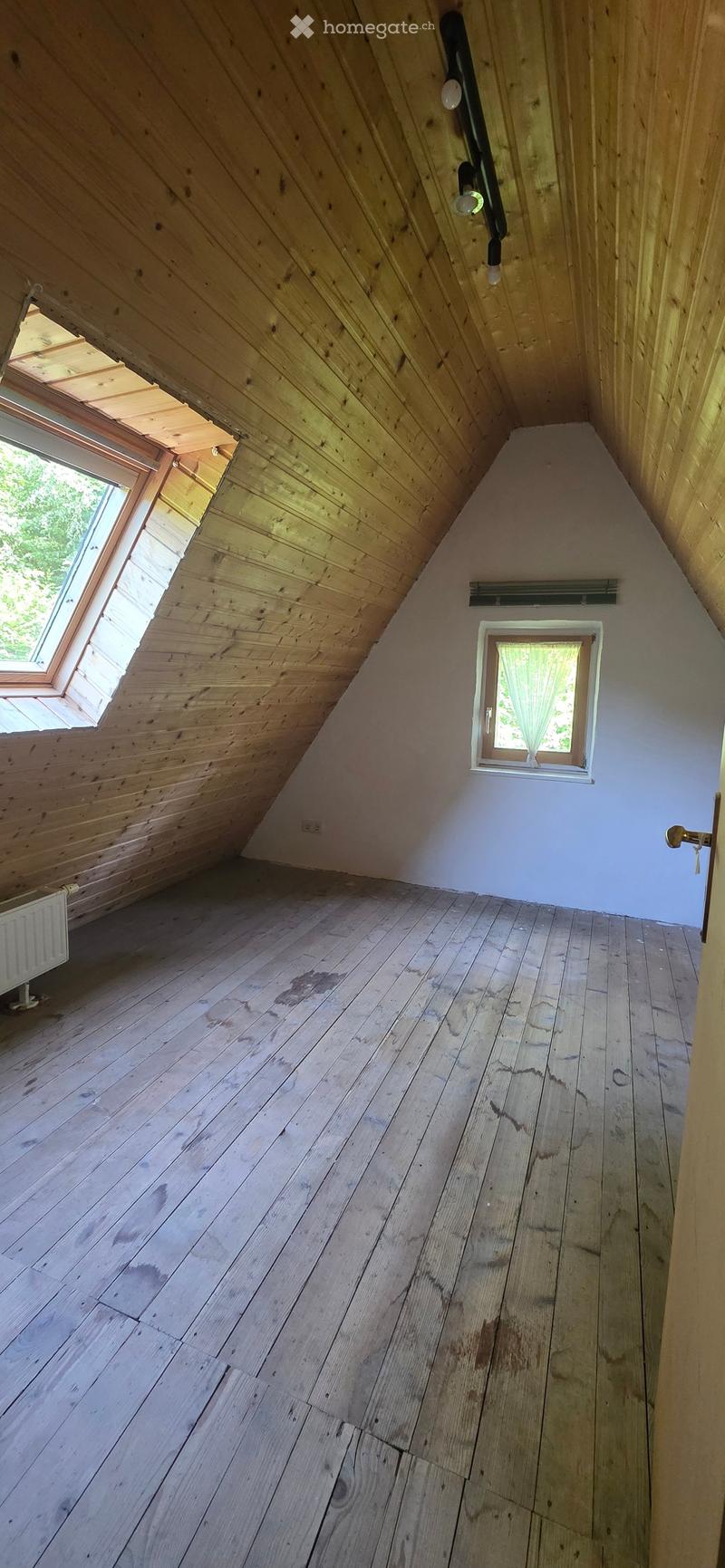 Empty attic room with wooden walls, wooden floor, skylight window, and small window with white frame.