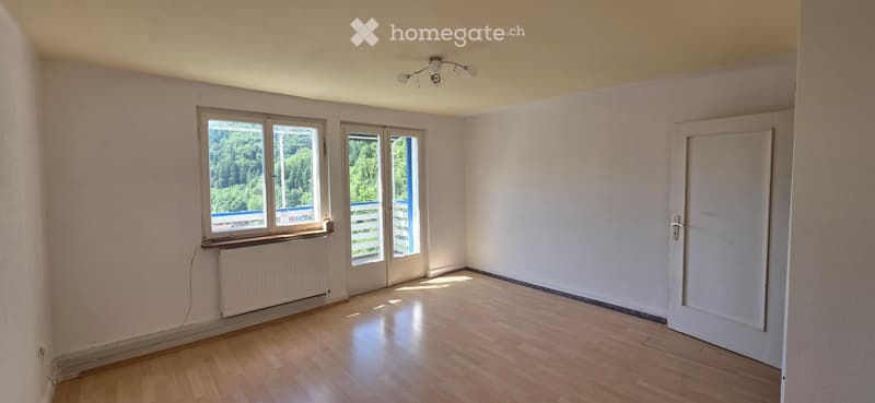 Empty room with wooden flooring, white walls, large windows with a view of greenery, and a radiator under one window.