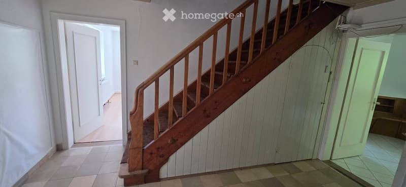 Wooden staircase with handrail leading to an upper level, white walls, tiled floor, and open doorway leading to another room.