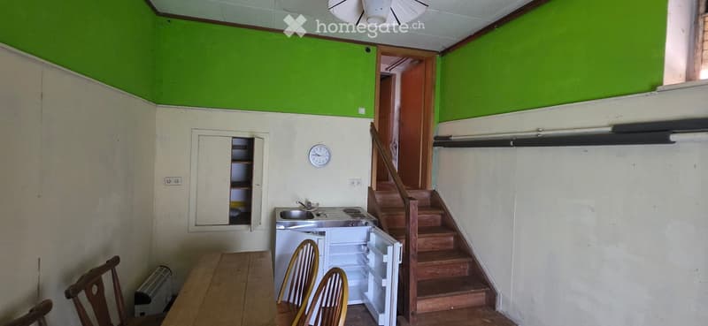 A small kitchen area with a wooden table, chairs, a small refrigerator, a sink, and a stove. There is a staircase leading upwards and a cabinet with shelves.