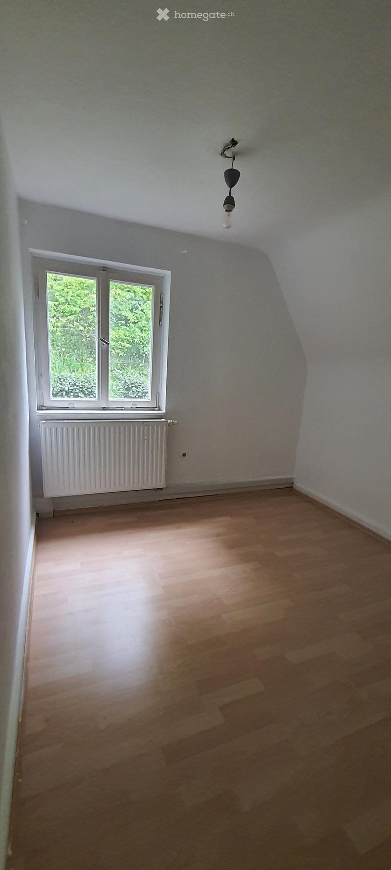 Empty room with wooden flooring, white walls, a single window with white frame, a radiator below the window, and a single hanging light fixture.