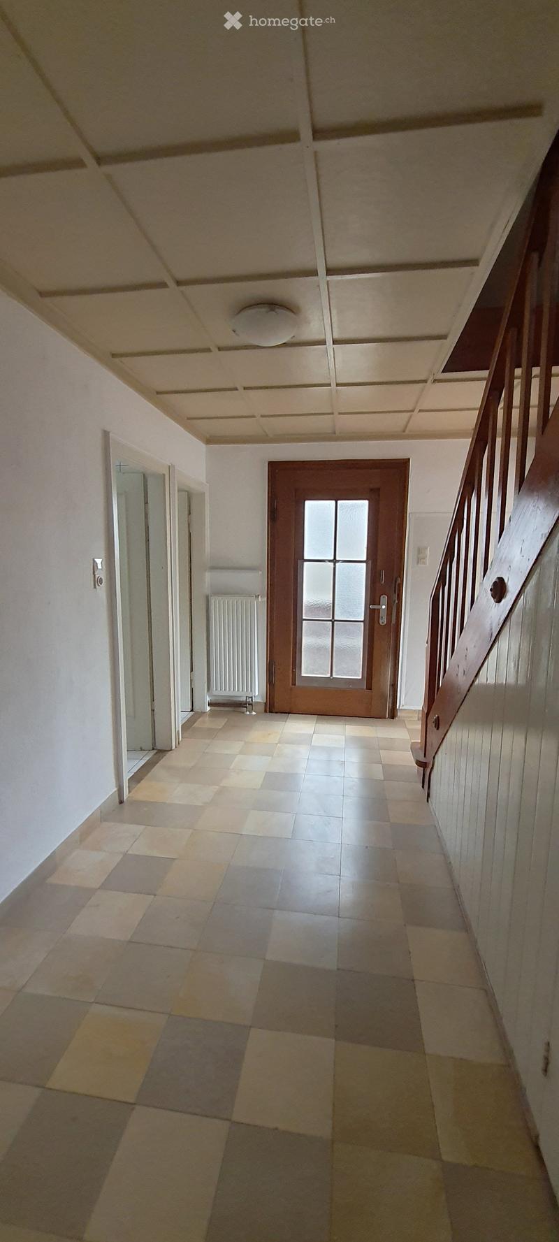 Empty hallway with tiled floor, white walls, a staircase on the right, and a wooden door at the end.