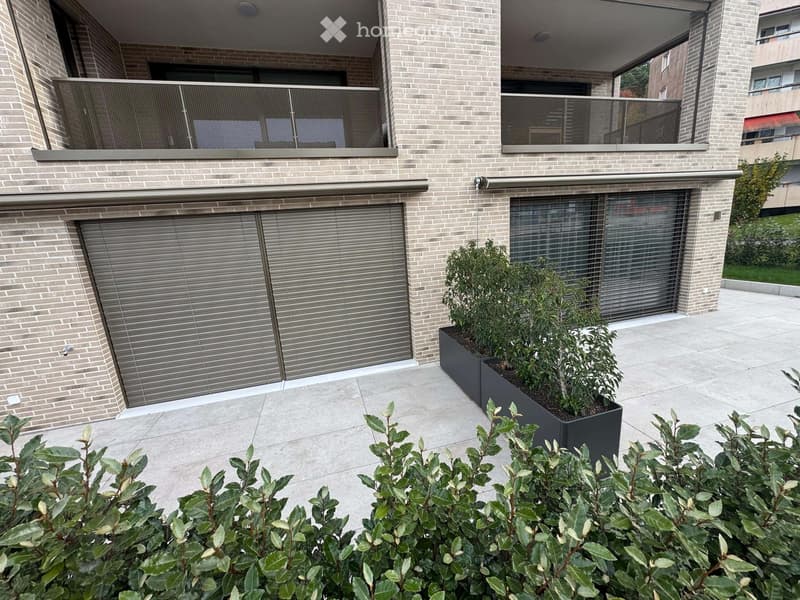 Modern outdoor area with a brick building facade, two large garage doors, raised planters with greenery, and a paved patio.