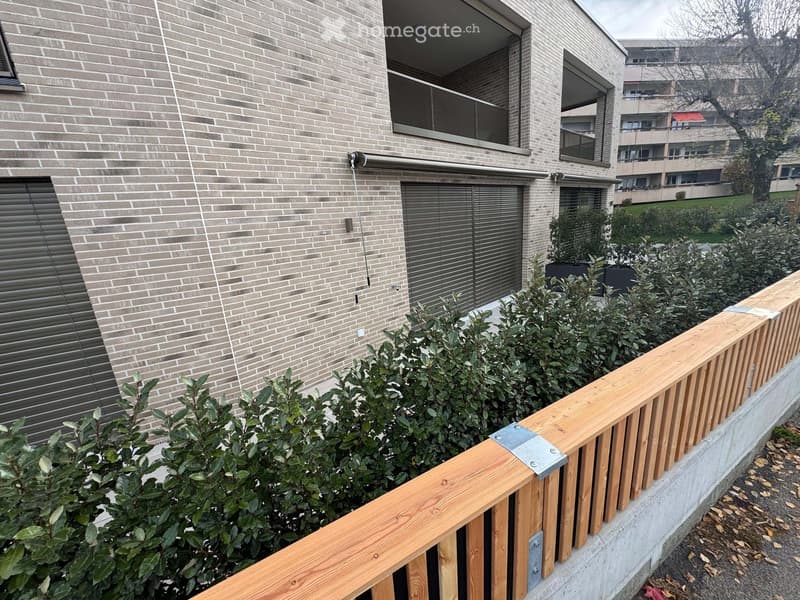 Modern building with brick facade, large windows, and balconies, surrounded by greenery and wooden railings.