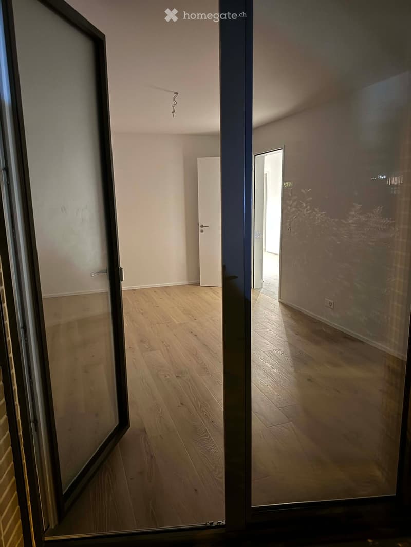 Empty hallway with light wooden flooring, white walls, and a closed door at the end. A glass sliding door is visible on the left side.