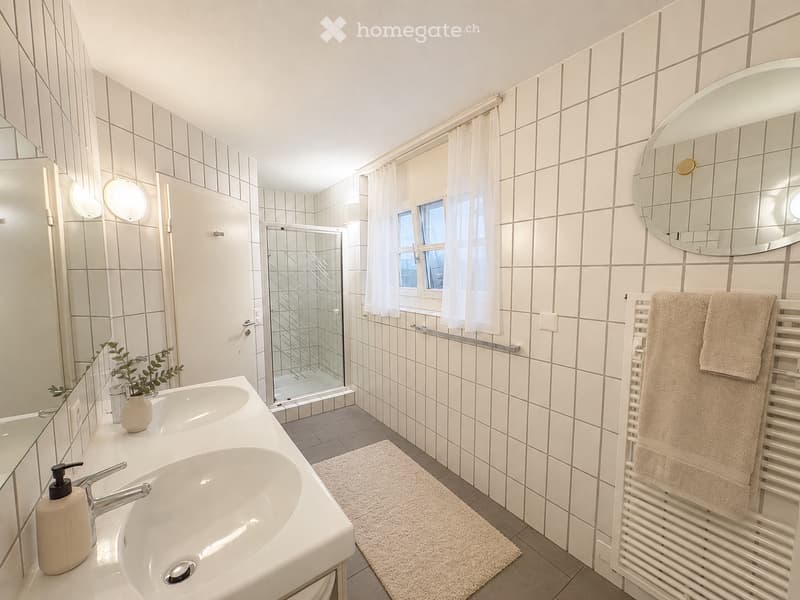 Modern bathroom with white tiled walls, a double sink, a large round mirror, a towel rack with towels, a shower area with a glass door, and a small plant on the sink counter.