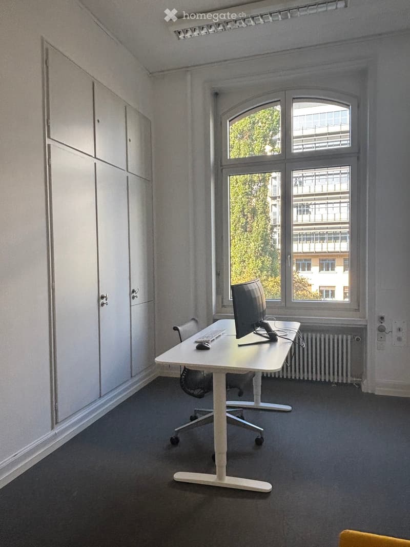 A small, well-lit room with a white desk, a computer, a chair, and a window offering a view of greenery and buildings.