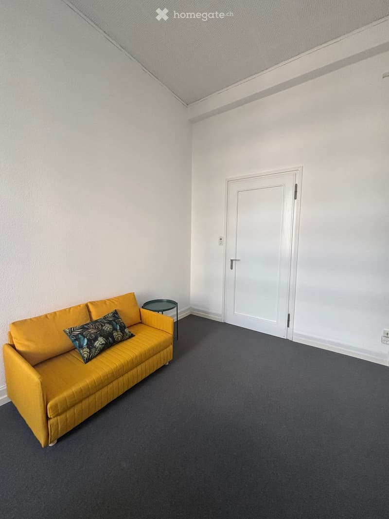 A small, furnished room with a yellow sofa, a small round table, and a decorative pillow. The room has white walls, a grey carpet, and a white door.