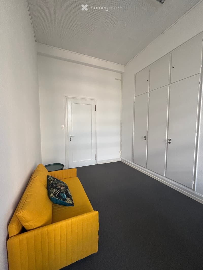 Bright, modern room with a yellow sofa, a decorative pillow, and a small round stool. The room features white walls, a dark grey carpet, and built-in white wardrobes.