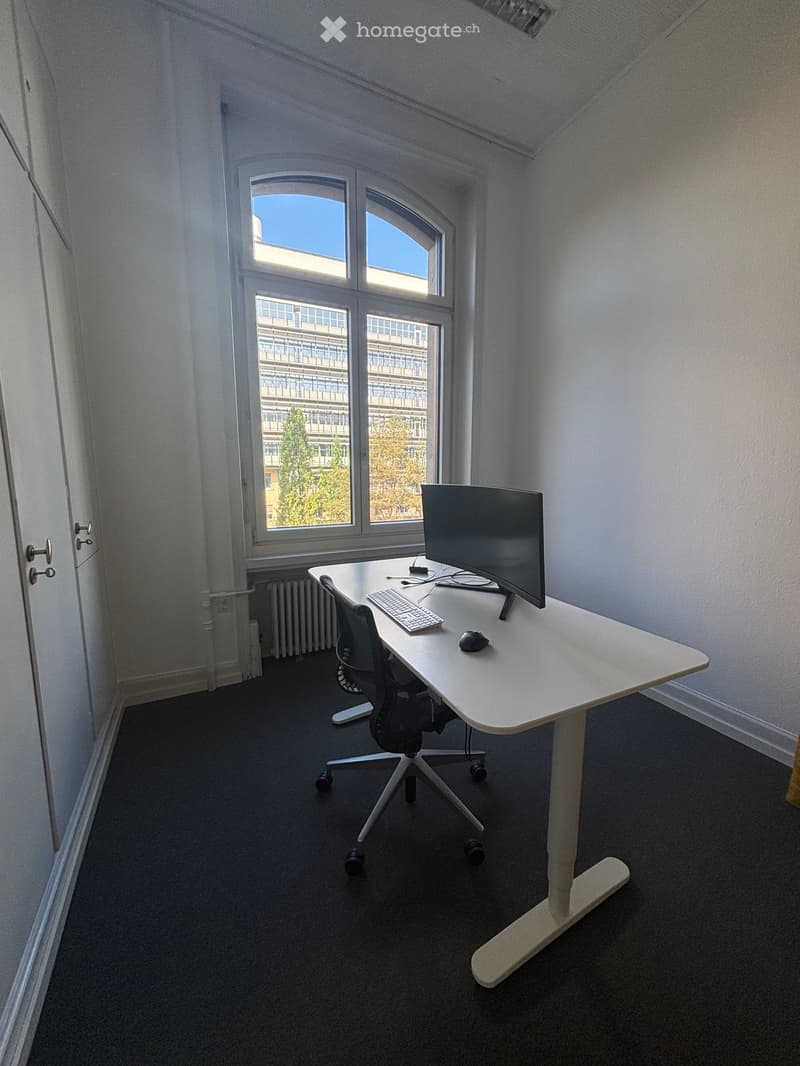 A modern, minimalist office space with a white desk, ergonomic chair, computer monitor, keyboard, and mouse. The room features large windows with a view of a cityscape and trees.
