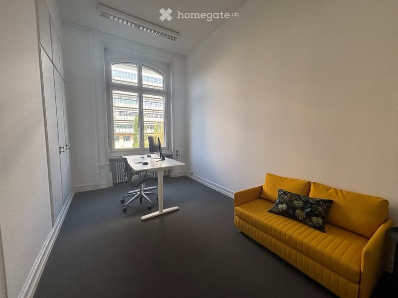 A modern, well-lit room with a yellow sofa, a desk with a computer, and a comfortable chair. Large windows provide natural light and a view of the outside.