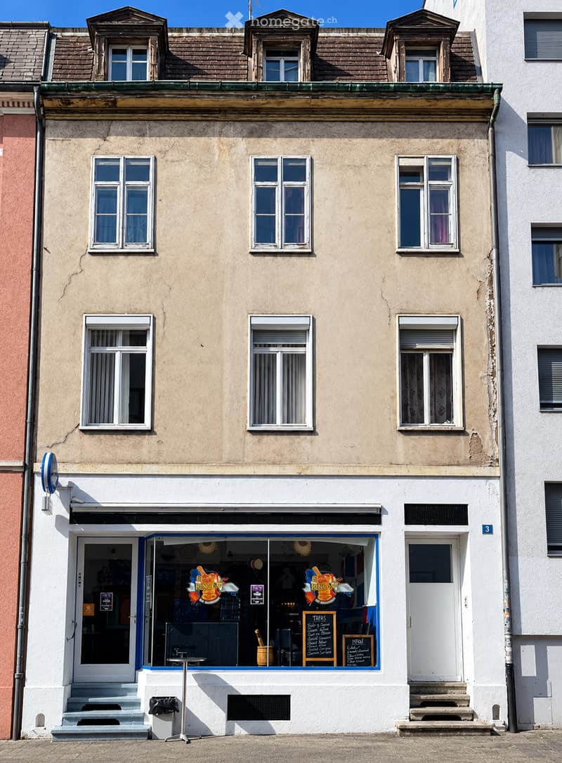 Three-story building with beige facade, multiple windows, and a storefront with signage reading "Rincon" and menu boards visible inside.