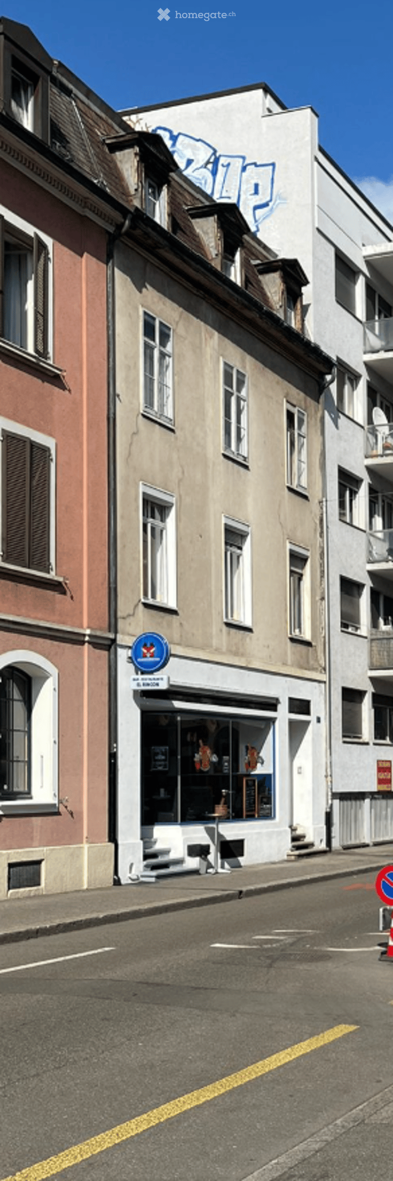 A row of multi-story buildings with varied facades, including a storefront with a sign and display window, and a painted mural on one building.