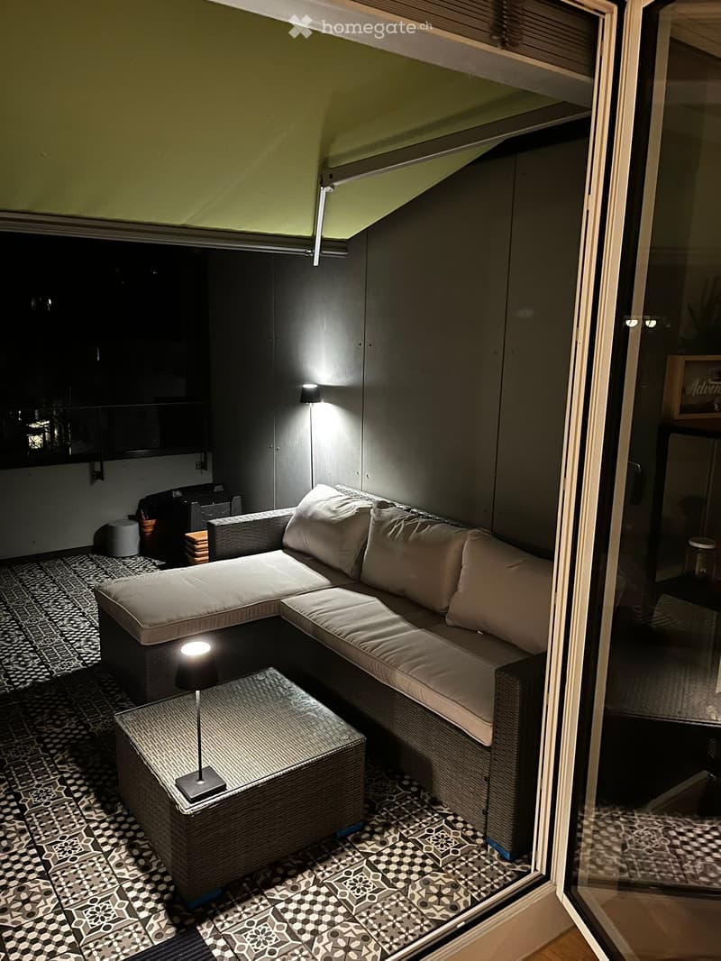 A modern, cozy living room with a grey sectional sofa, white cushions, a wicker coffee table, and a patterned tile floor.