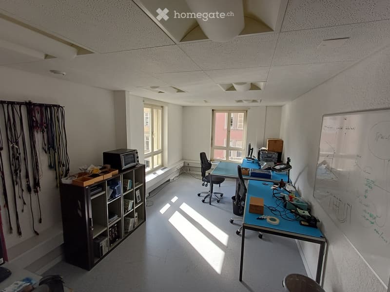 Office space with two desks, chairs, a shelf with equipment, a microwave, and a whiteboard. Natural light streams in from large windows.