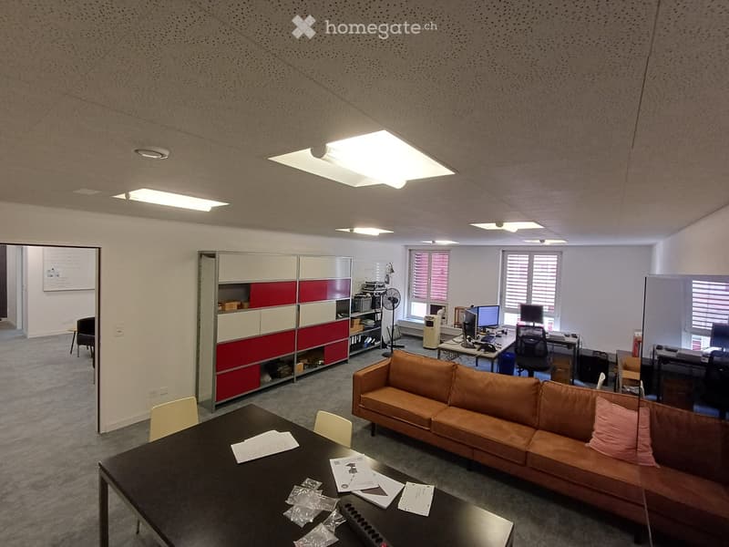 Office space with a brown leather sofa, black table with papers, white chairs, computer screens, and a red and white cabinet.