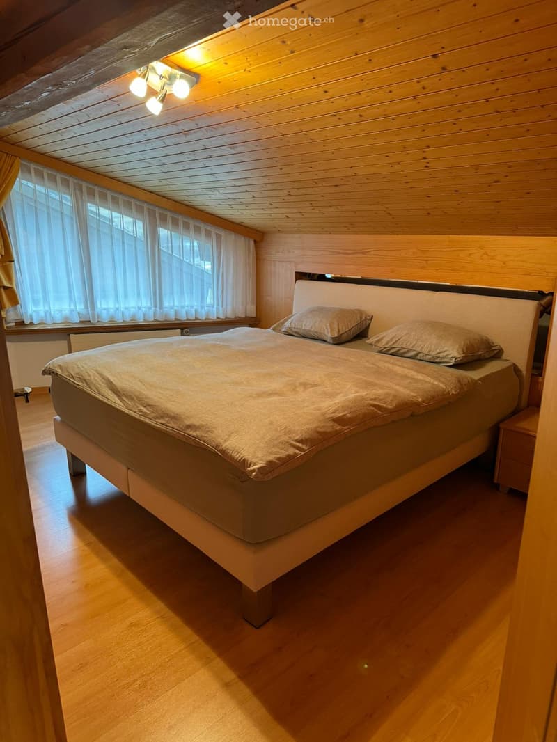 Bedroom with wooden floor, wooden ceiling, large bed, two pillows, nightstand, large window with white curtains