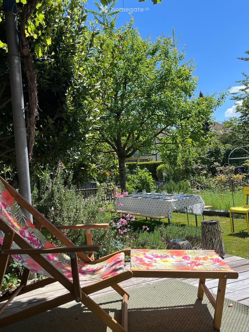 Garden with a wooden deck, a table with a white tablecloth, chairs, a bench, and a small stool, surrounded by greenery and flowers.