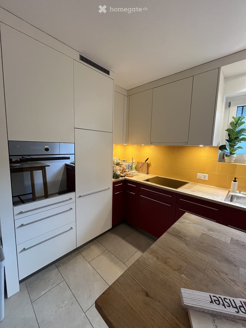 Modern kitchen with white and dark red cabinets, a central island, and a built-in stove. Features include a sink, a wooden table, and a potted plant on the windowsill.