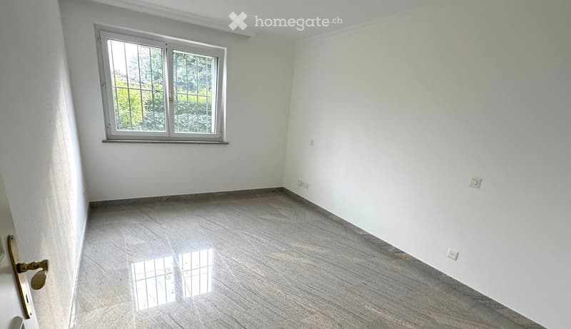 Empty room with white walls, grey tiled floor, and a window with metal bars. No furniture or equipment present.